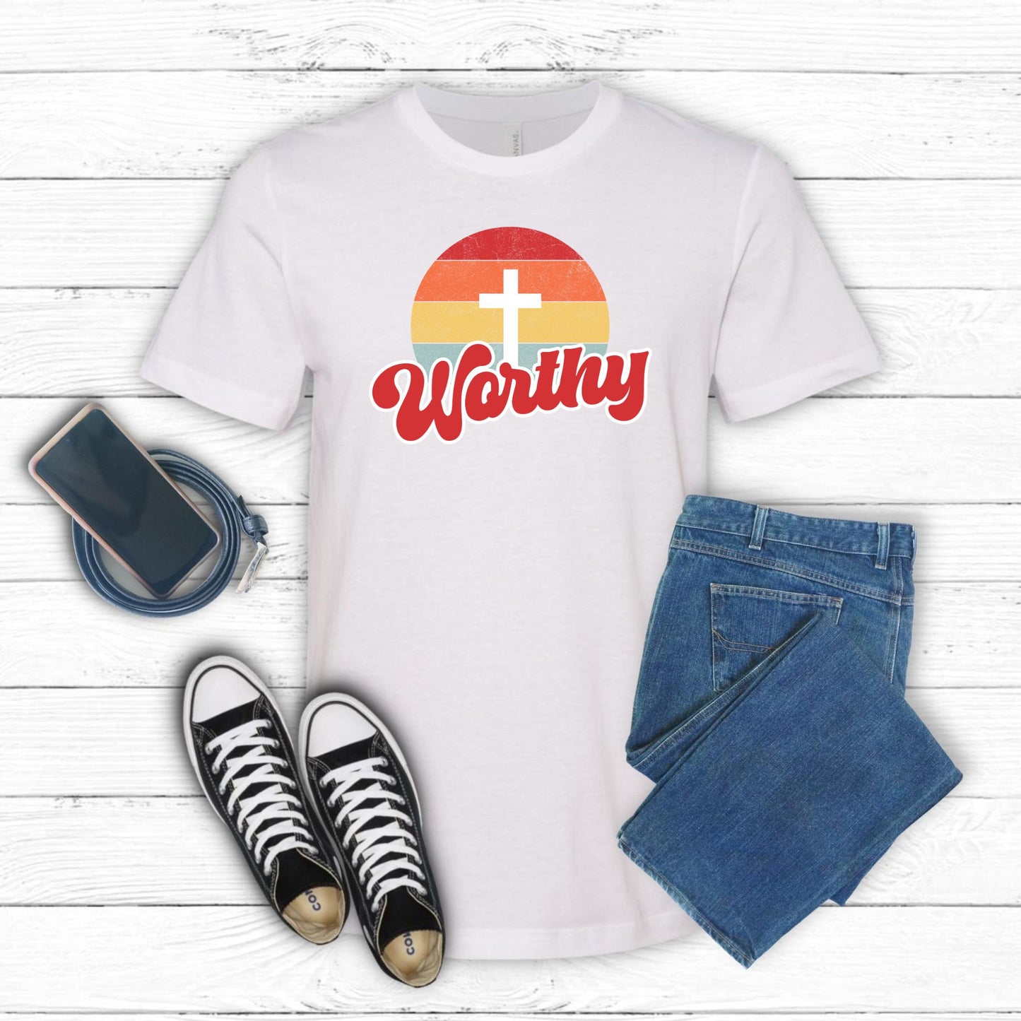 White t-shirt with 'Worthy' design, black sneakers, blue jeans, and phone on a white wooden surface