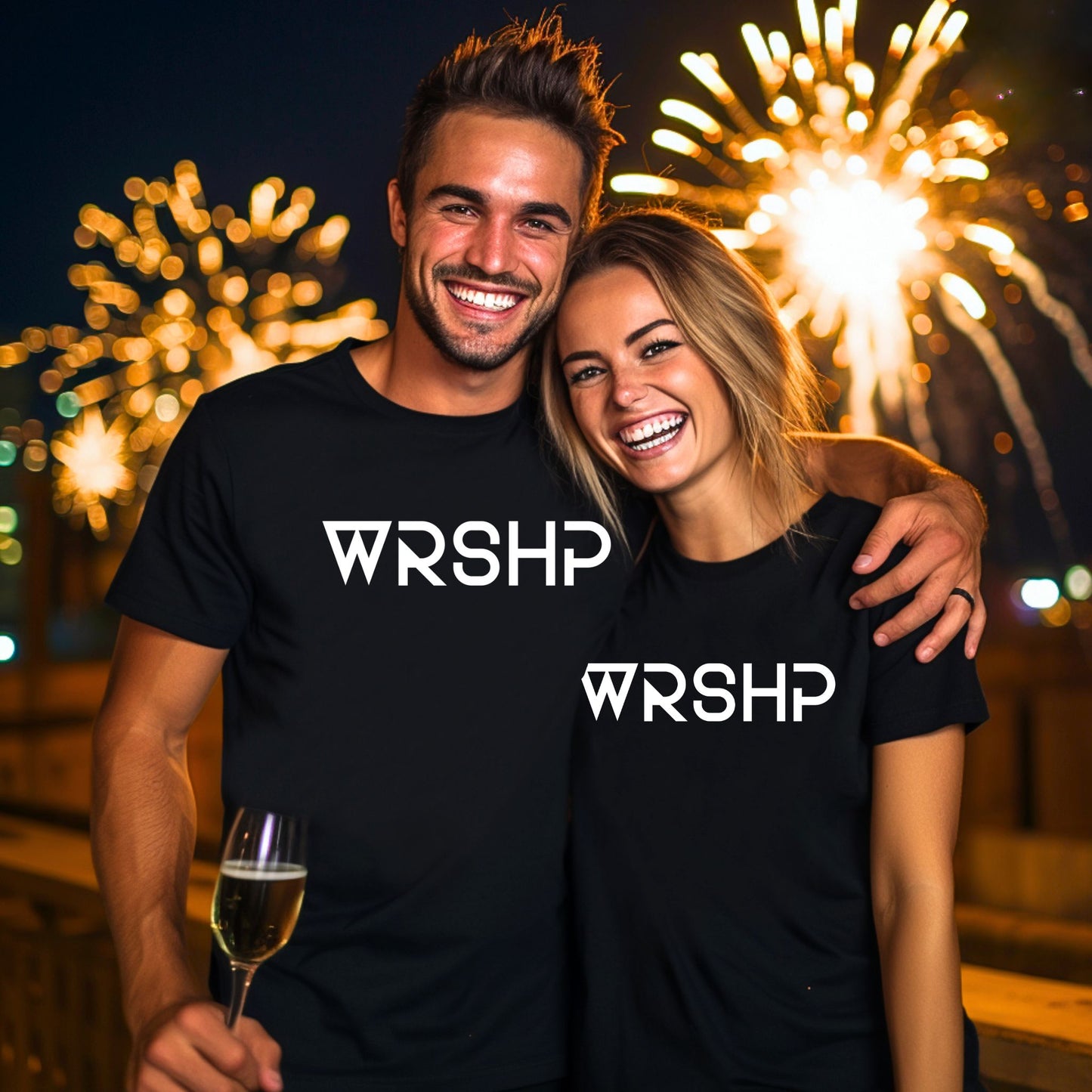 Two people wearing black t-shirts with 'WRSHP' logo, smiling in front of fireworks.
