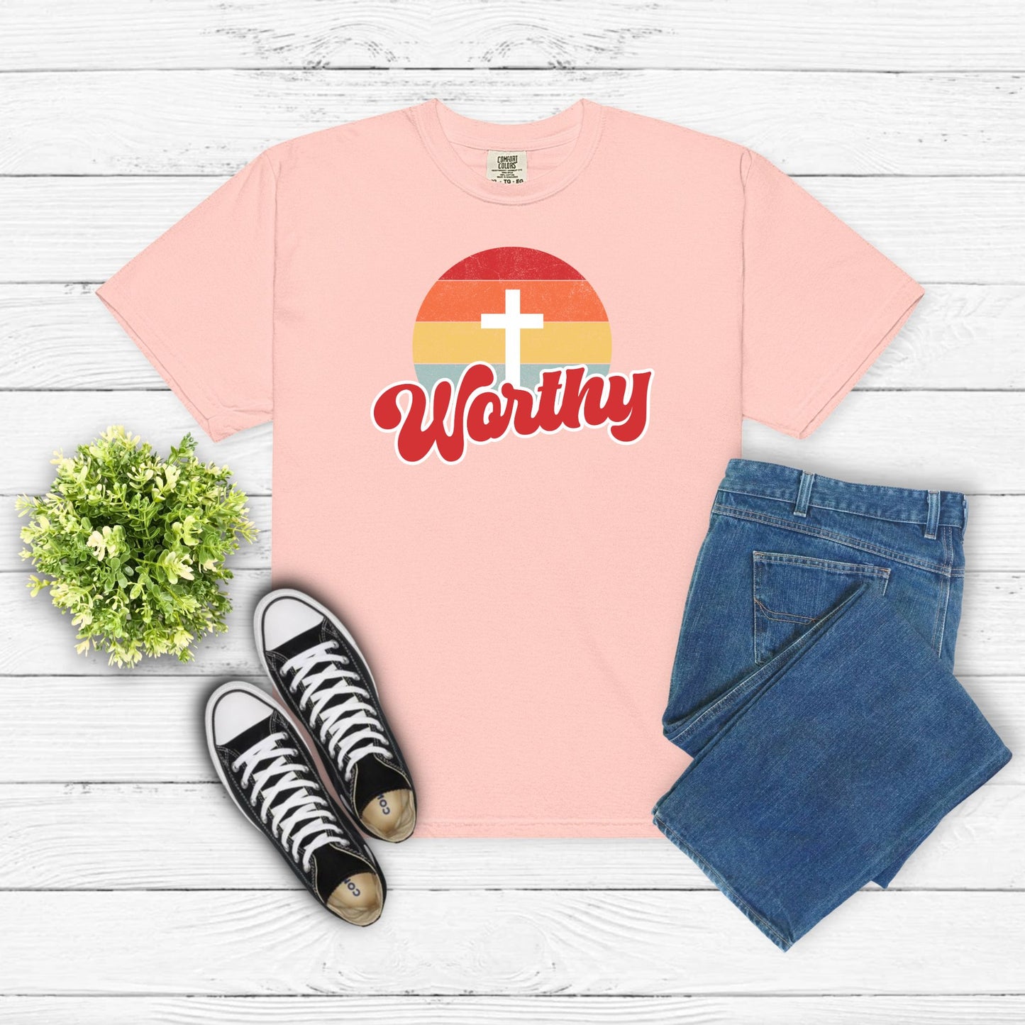 Pink t-shirt with 'Worthy' design, black sneakers, and blue jeans on a wooden surface.